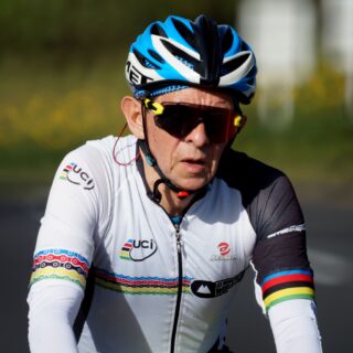 Cyclist wearing a blue helmet and red-tinted sunglasses, white racing jersey with sponsor logos, outdoors.