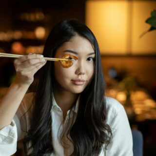 Woman with long dark hair in a white shirt holds a shrimp on chopsticks up to her eye in a warmly lit restaurant setting in the background.