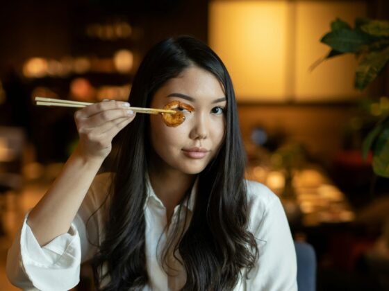 Woman with long dark hair in a white shirt holds a shrimp on chopsticks up to her eye in a warmly lit restaurant setting in the background.