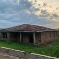 Rusted corrugated metal roof on a brick building with an open entrance, green lawn in the foreground, cloudy sky overhead at dusk.