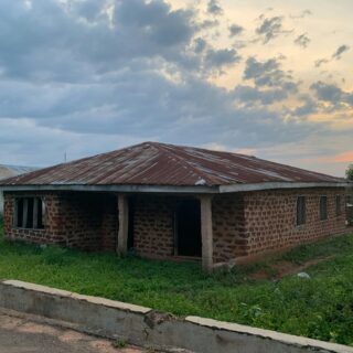 Rusted corrugated metal roof on a brick building with an open entrance, green lawn in the foreground, cloudy sky overhead at dusk.