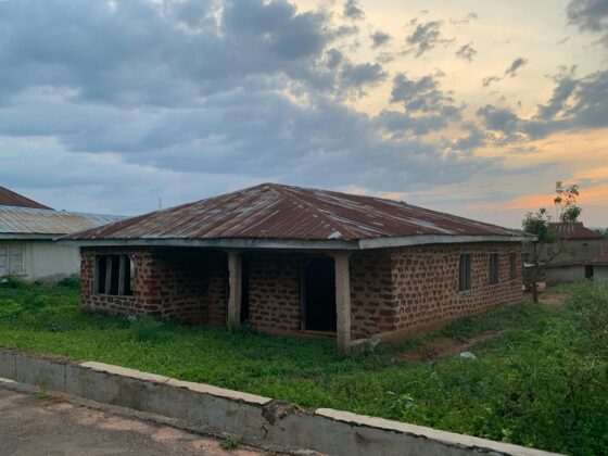 Rusted corrugated metal roof on a brick building with an open entrance, green lawn in the foreground, cloudy sky overhead at dusk.