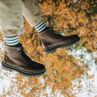 Person wearing brown lace-up boots stepping through fallen autumn leaves with green-and-white striped socks visible above the boots