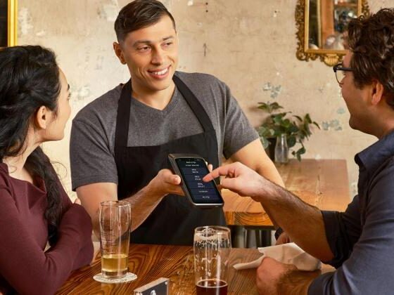 Server in a gray shirt and black apron shows a mobile payment device to a couple at a wooden table with drinks.
