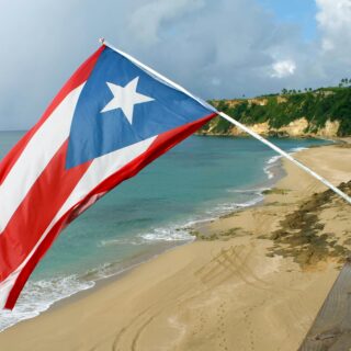 Puerto Rico flag waving on a pole beside a tropical beach with turquoise water and rocky cliffs in the background.