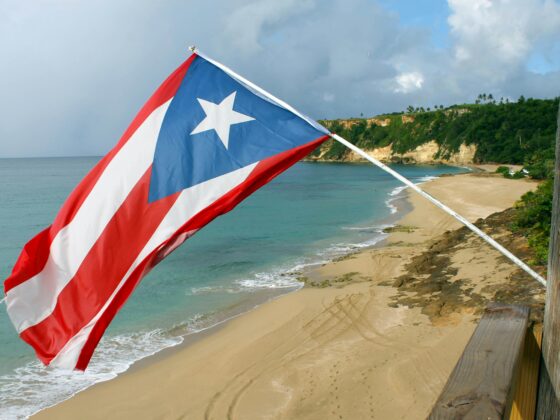 Puerto Rico flag waving on a pole beside a tropical beach with turquoise water and rocky cliffs in the background.