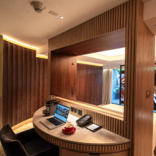 Curved wooden desk with a laptop and red cup in a luxury hotel suite, warm lighting and a mirror overlooking the bed area.