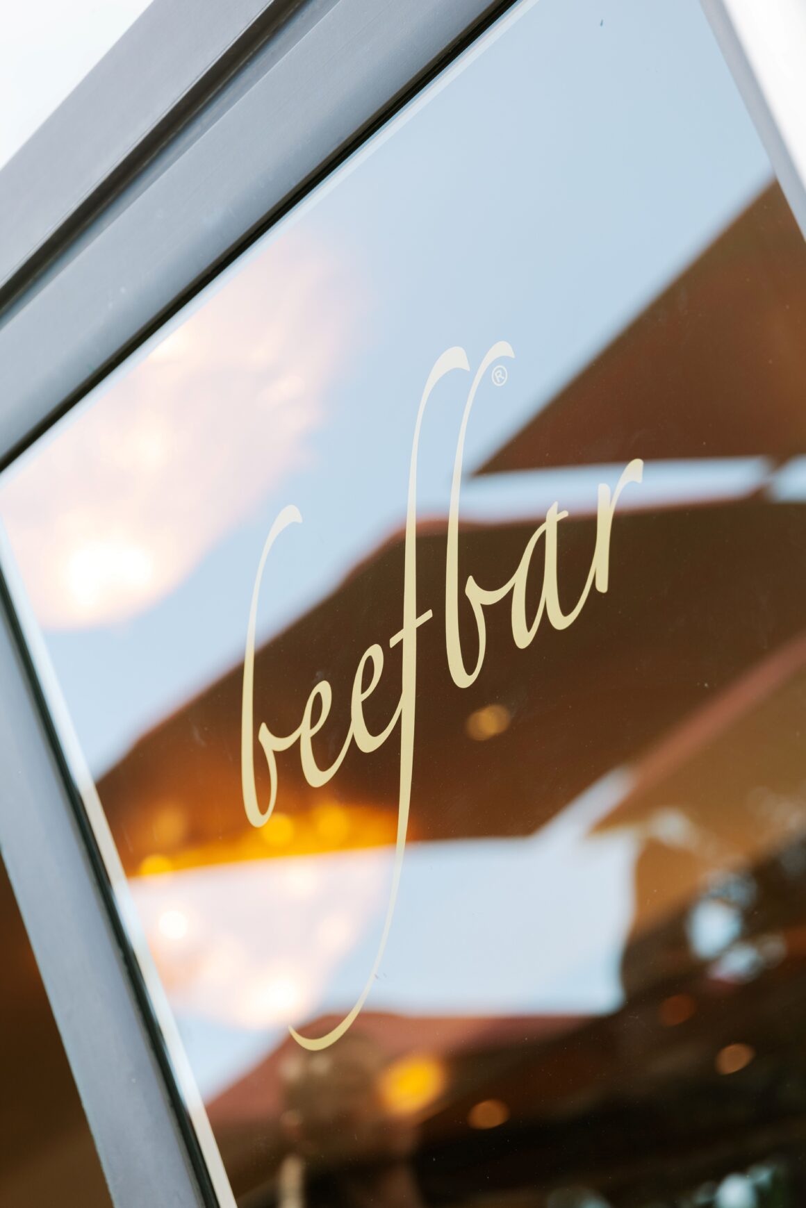 Close-up of a glass window with pale yellow cursive lettering and a metal frame, reflecting the sky.