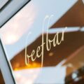 Close-up of a glass window with pale yellow cursive lettering and a metal frame, reflecting the sky.