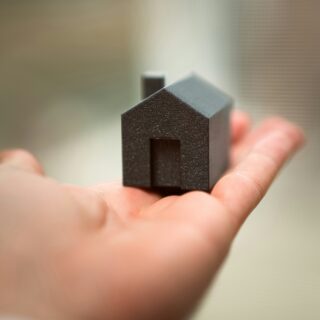 Close-up of a tiny black house-shaped model held between two fingers with a blurred background on a neutral wall/desk surface.