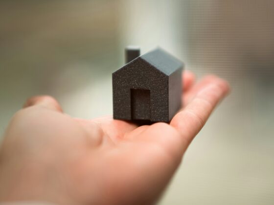 Close-up of a tiny black house-shaped model held between two fingers with a blurred background on a neutral wall/desk surface.
