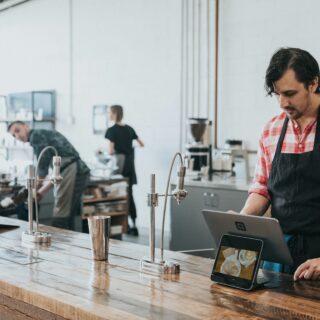 Barista in a red plaid shirt and black apron uses a tablet POS at a wooden cafe counter.
