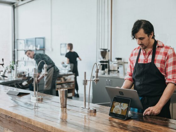 Barista in a red plaid shirt and black apron uses a tablet POS at a wooden cafe counter.