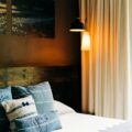 Bed with blue-striped pillows on white linens, a wall photo, and warm lamps beside beige curtains for a cozy bedroom scene.