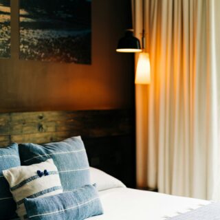 Bed with blue-striped pillows on white linens, a wall photo, and warm lamps beside beige curtains for a cozy bedroom scene.
