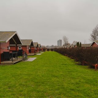 Row of red wooden cabins with gray roofs along a green lawn on an overcast day, a bicycle resting near a patio table in the foreground.