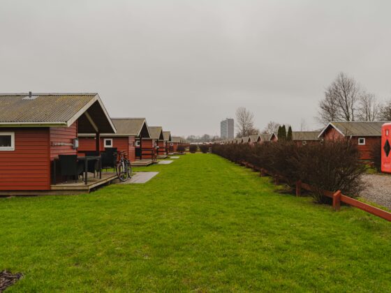 Row of red wooden cabins with gray roofs along a green lawn on an overcast day, a bicycle resting near a patio table in the foreground.