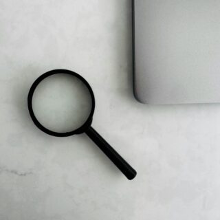 Close-up of a black magnifying glass resting on a light, marble-like desk near the corner of a silver laptop edge.