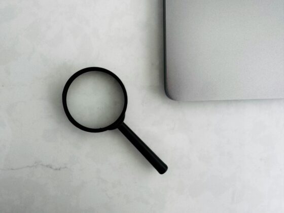 Close-up of a black magnifying glass resting on a light, marble-like desk near the corner of a silver laptop edge.