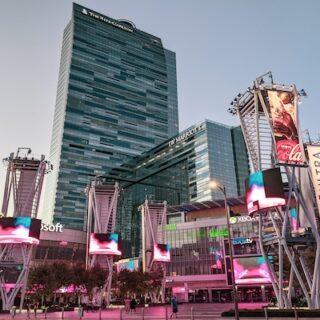 Busy city square in front of tall glass skyscrapers with large digital billboards and pink lighting at dusk.