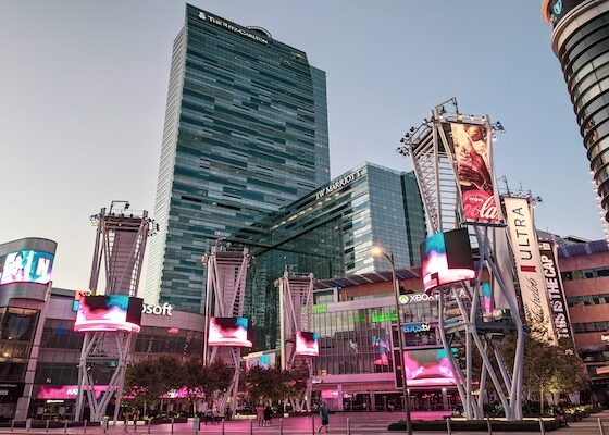 Busy city square in front of tall glass skyscrapers with large digital billboards and pink lighting at dusk.