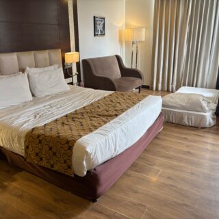 Hotel room with two large beds, beige headboard, and patterned runners; a brown chair and floor lamp beside the window curtains.