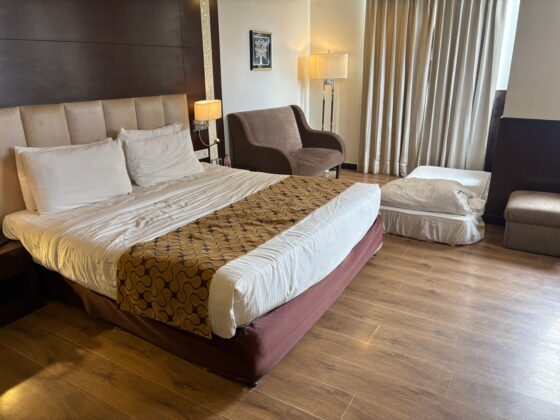Hotel room with two large beds, beige headboard, and patterned runners; a brown chair and floor lamp beside the window curtains.