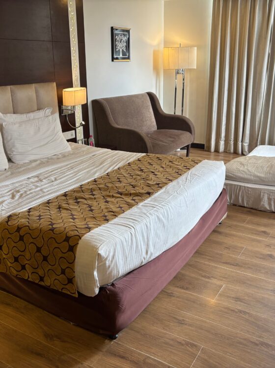 Hotel room with two large beds, beige headboard, and patterned runners; a brown chair and floor lamp beside the window curtains.
