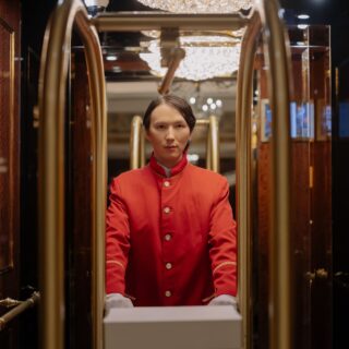 Porter in a red uniform standing inside a luxurious elevator with wood walls and crystal chandeliers in the ceiling.