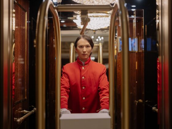 Porter in a red uniform standing inside a luxurious elevator with wood walls and crystal chandeliers in the ceiling.
