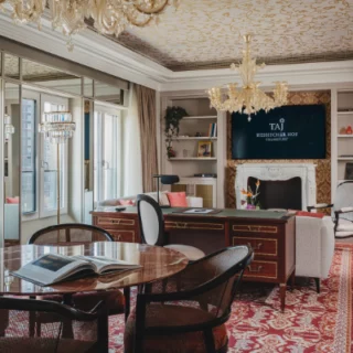 Luxurious hotel executive office with a round marble table and wicker chairs, a fireplace, chandeliers, built-in bookshelves, and a TV displaying the Taj logo on the wall.