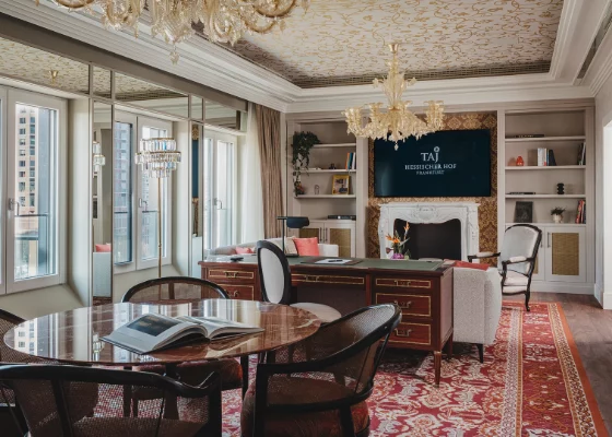 Luxurious hotel executive office with a round marble table and wicker chairs, a fireplace, chandeliers, built-in bookshelves, and a TV displaying the Taj logo on the wall.