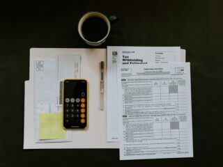 Top-down view of a workspace with a black coffee mug, calculator, pen, sticky notes, and tax documents on a white desk.