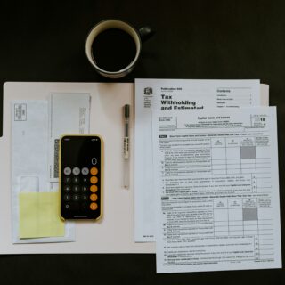 Top-down view of a workspace with a black coffee mug, calculator, pen, sticky notes, and tax documents on a white desk.