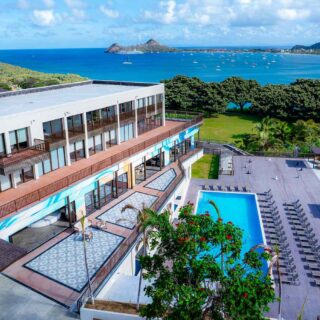 Aerial view of a seaside resort with a rectangular pool, lounge chairs, and a white building overlooking a blue bay.