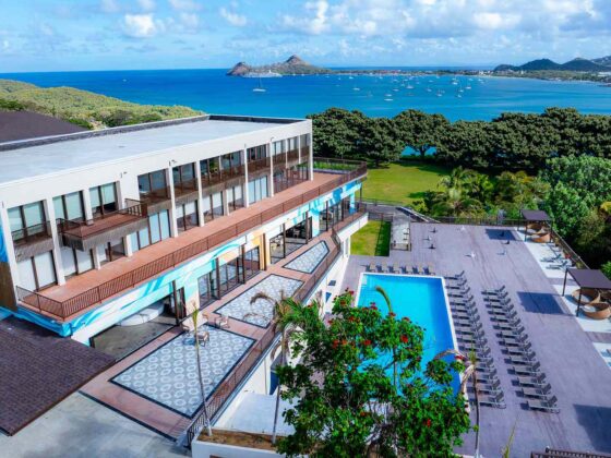 Aerial view of a seaside resort with a rectangular pool, lounge chairs, and a white building overlooking a blue bay.