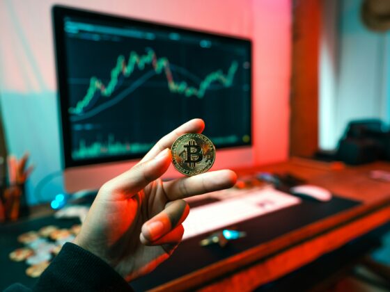 Person holds a gold Bitcoin coin up to the camera with a computer monitor displaying a fluctuating stock chart in the background under colorful lighting