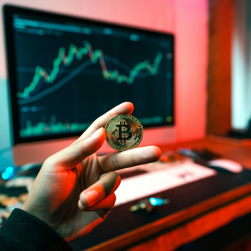 Person holds a gold Bitcoin coin up to the camera with a computer monitor displaying a fluctuating stock chart in the background under colorful lighting