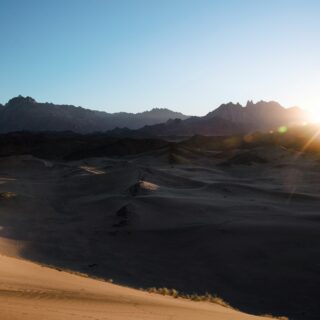 Hiker descends a sandy dune in a desert with dark mountains in the distance as the sun sets on the right.