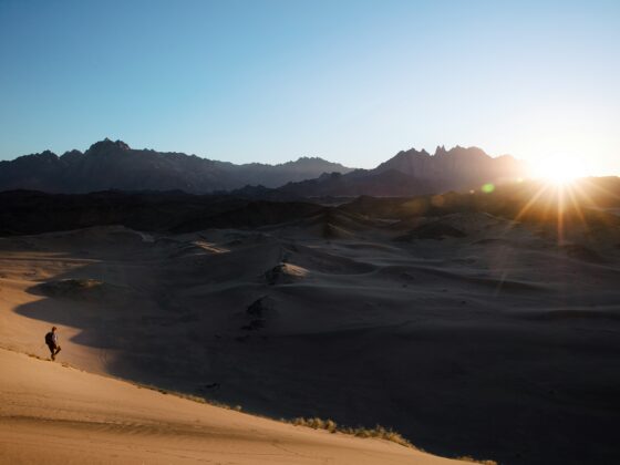 Hiker descends a sandy dune in a desert with dark mountains in the distance as the sun sets on the right.