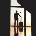Silhouette of a traveler with a rolling suitcase standing in a dim terminal, gazing at an airplane in the sunset outside the window.