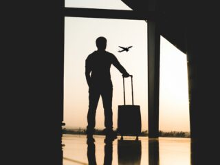 Silhouette of a traveler with a rolling suitcase standing in a dim terminal, gazing at an airplane in the sunset outside the window.