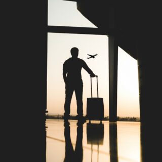Silhouette of a traveler with a rolling suitcase standing in a dim terminal, gazing at an airplane in the sunset outside the window.