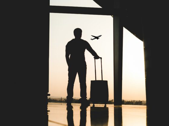 Silhouette of a traveler with a rolling suitcase standing in a dim terminal, gazing at an airplane in the sunset outside the window.