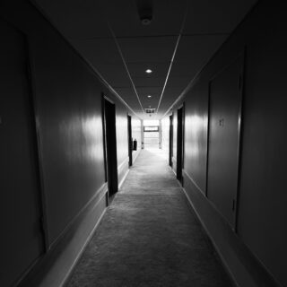 Dim, black-and-white hotel hallway stretching to a bright exit at the end, doors along both sides.