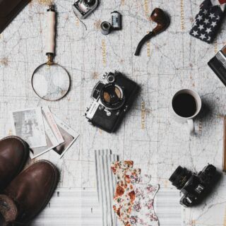 Overhead view of travel gear laid out on a map: vintage camera, magnifying glass, film canister, boots, coffee mug, and patterned fabric.