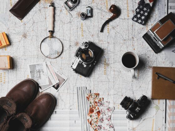 Overhead view of travel gear laid out on a map: vintage camera, magnifying glass, film canister, boots, coffee mug, and patterned fabric.