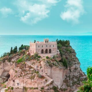 Beige castle-like building perched on a rocky cliff above a blue sea, with palm trees and a car-filled road at the base.