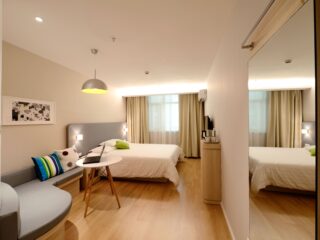 Hotel bedroom with a white bed, gray headboard, colorful pillows, a small round table with a laptop, and a gray sofa in a bright, modern space.