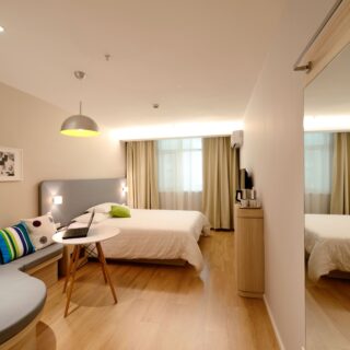 Hotel bedroom with a white bed, gray headboard, colorful pillows, a small round table with a laptop, and a gray sofa in a bright, modern space.